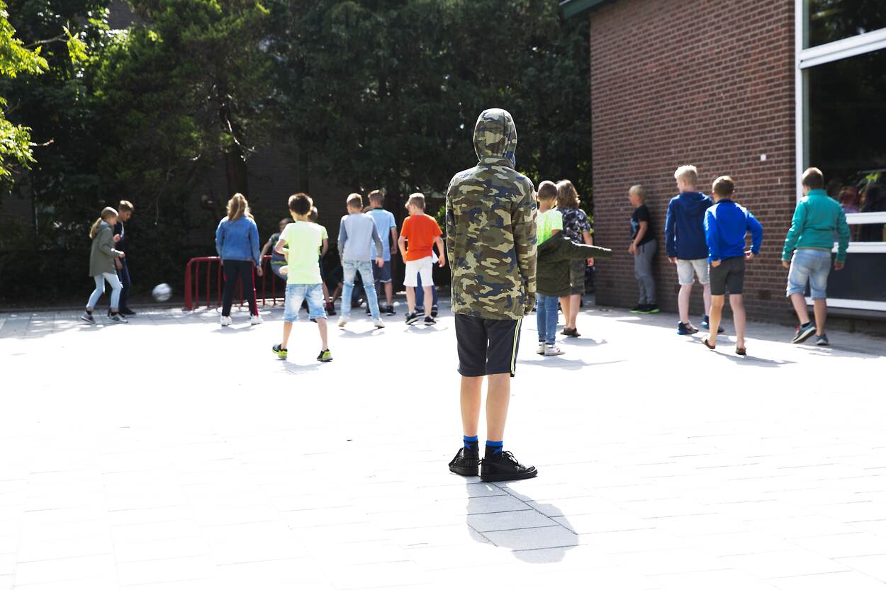 Op een stenen plein wordt door jongens en meisjes gevoetbald. Een kind met een legerjas aan, een capuchon op en een korte broek aan kijkt toe.