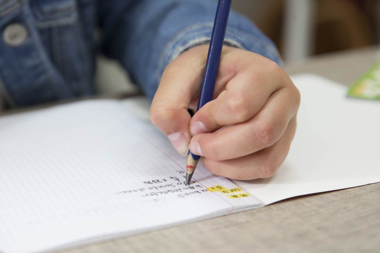 In een schrift wordt met een potlood een antwoord opgeschreven. Het kind draagt een spijkerjas en staat niet volledig op beeld. Enkel met één hand.