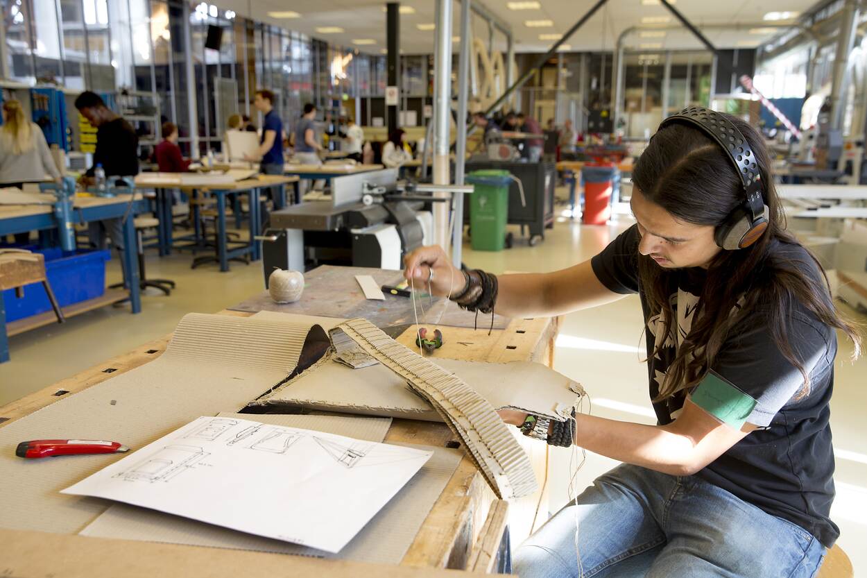 In een technieklokaal wordt gewerkt door verschillende jongeren. Eén jongen met een koptelefoon op werkt aan een ontwerp met karton en touw.