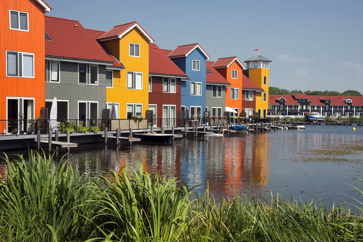 Aan het water staan gekleurde huizen met een steiger op het water. Aan sommige steigers ligt een bootje. Op een steiger zit een vrouw in bikini.