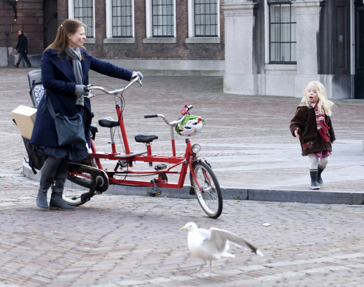 Een vrouw heeft een tandemfiets vast. Aan het voorste stuur hangt een kinderhelm. Naast de tandem staat een klein meisje die naar een meeuw kijkt.