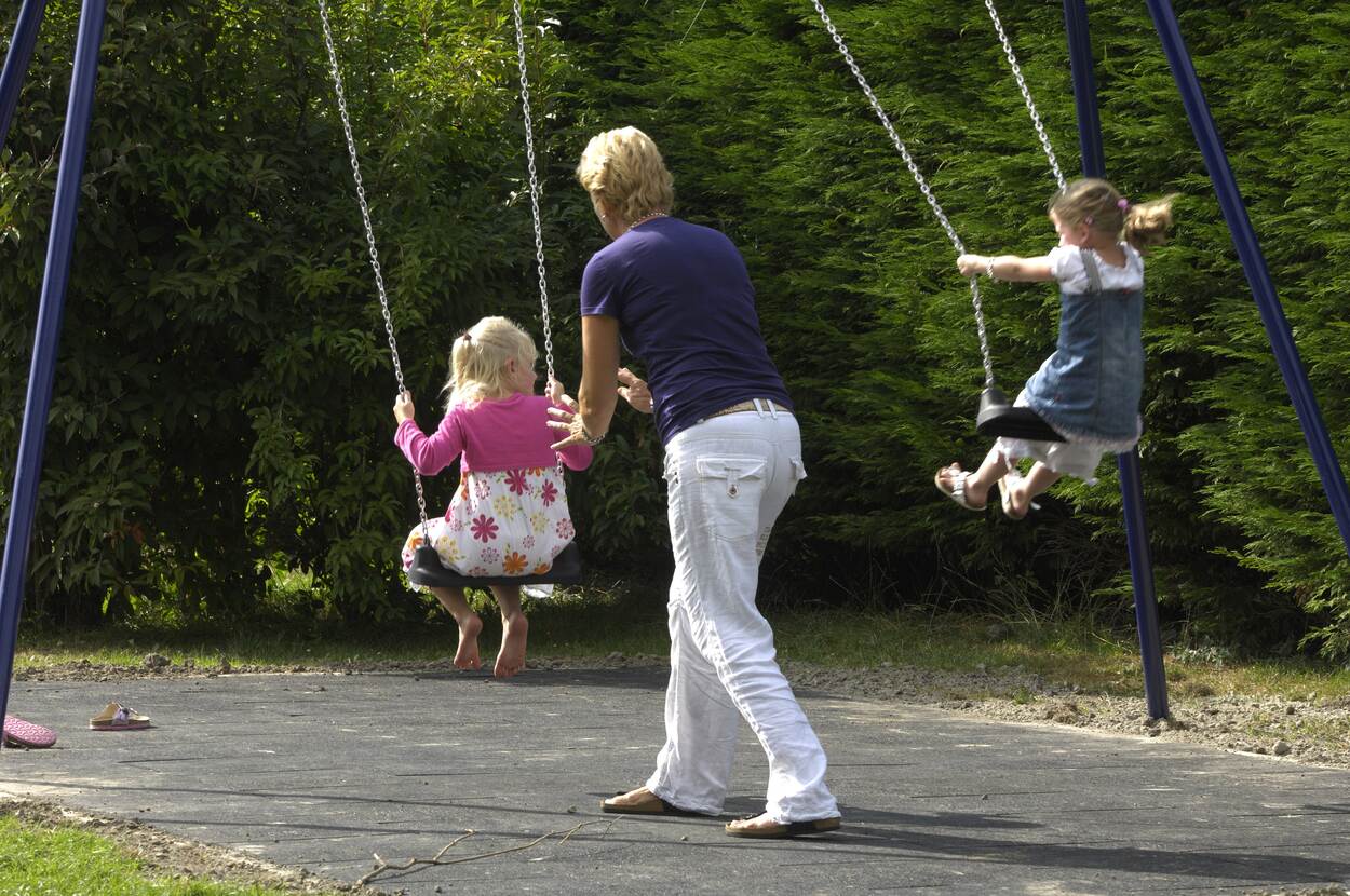 Twee meisjes zijn aan het schommelen. Een meisje een bloemetjes jurk en roze vestje wordt geduwd door een vrouw. Het andere meisje schommelt zelf.