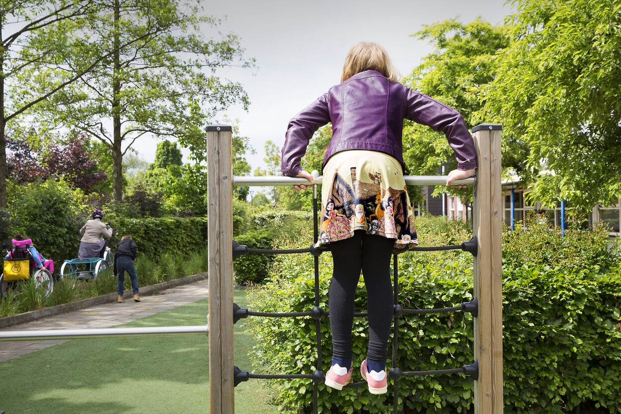 Een meisje klimt op een klimrek. Links van het meisje gaat iemand in een rolstoel en een aangepaste fiets voorbij. Een jongen loopt langs.