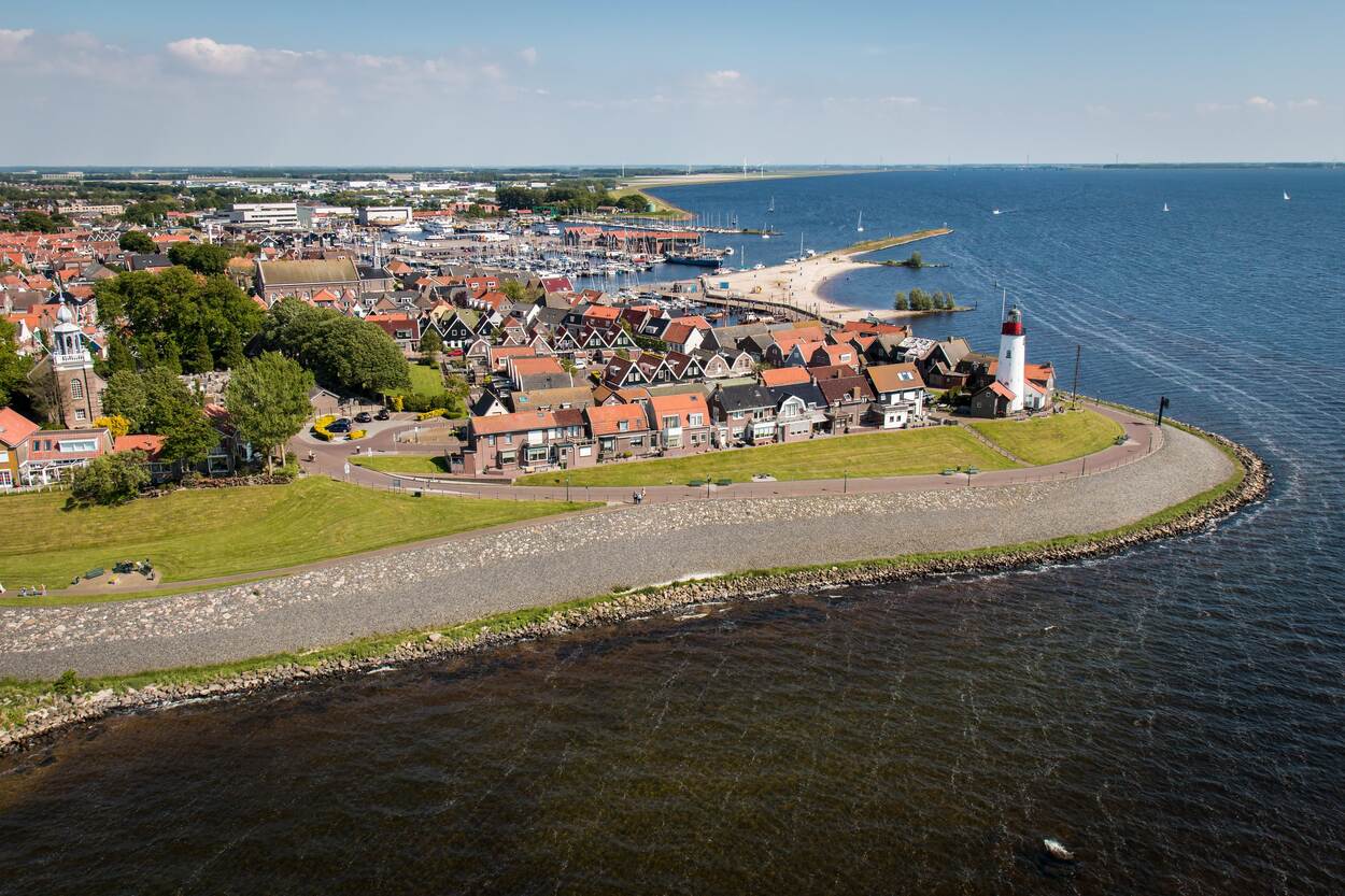 Vanuit de lucht kijk je uit op de gemeente Urk. Je ziet aan het water de dijk met een vuurtoren en de haven met schepen. Achter de dijk staan huizen.