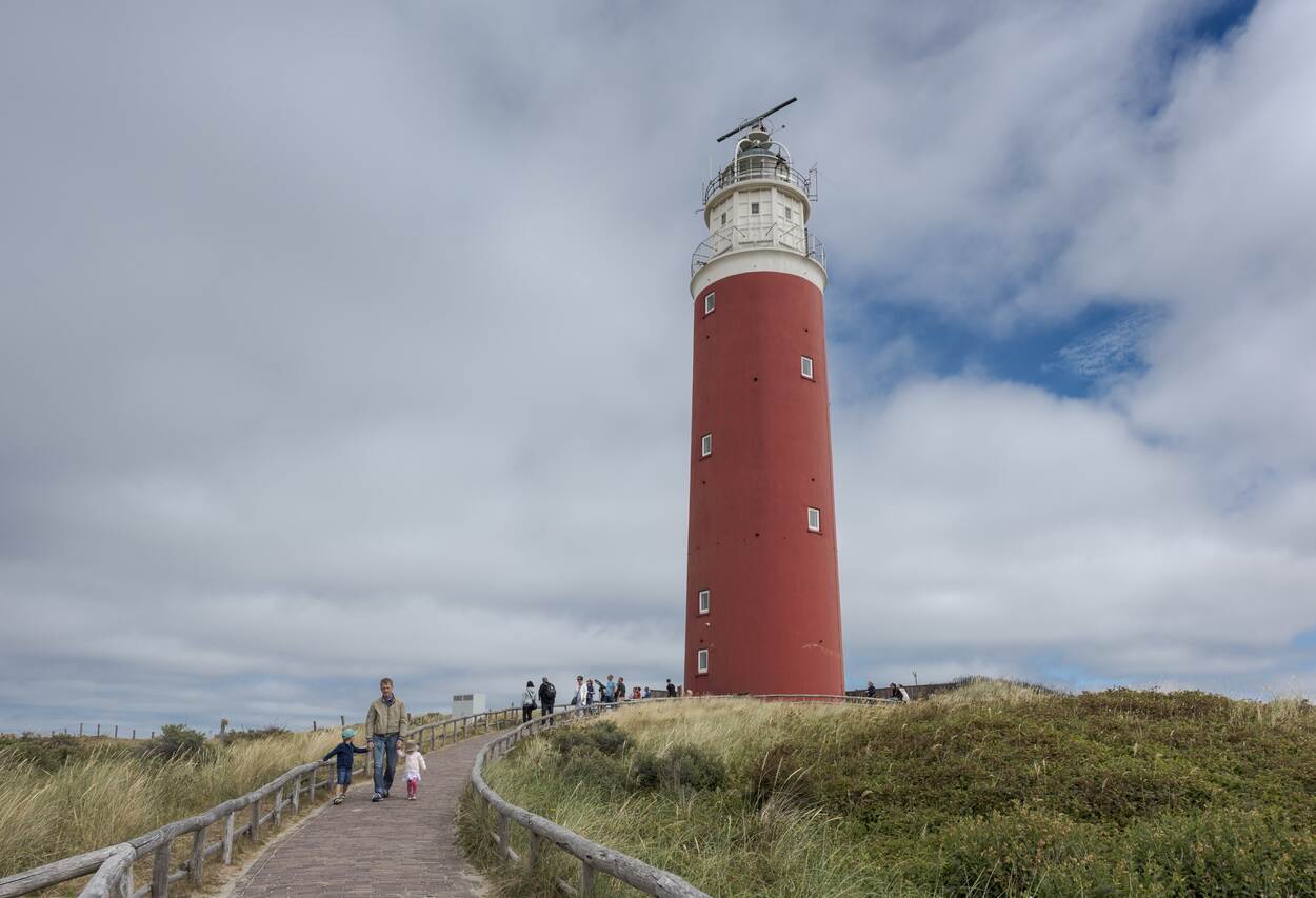 Een vuurtoren in de duinen van Texel met op de achtergrond witte wolken met hier en daar wat blauwe lucht. Op het pad naar de vuurtoren lopen mensen