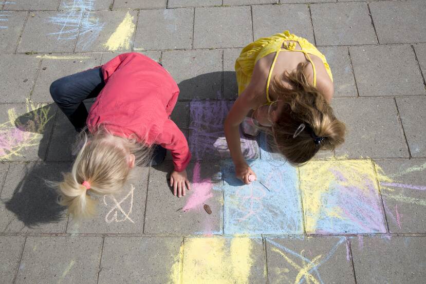 Eén meisje tekent met roze krijt een vlinder naast een roze hartje op een blauw gekrijte stoeptegel. Het andere meisje tekent iets gele krijt.