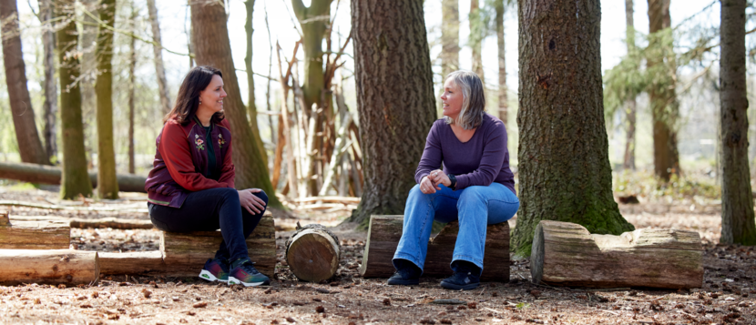 Astrid van Lier en Ilona van der Heijden zitten op boomstammen met een bos op de achtegrond.