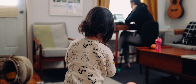 Een klein kind in een kamer met een volwassene achter de computer op de achtergrond.