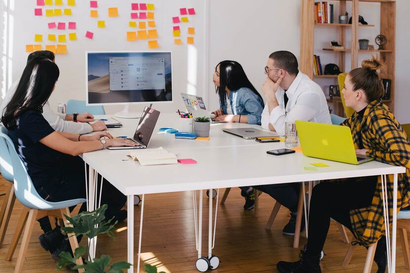 Een foto van een groep mensen die aan een tafel zit, sommigen van hen hebben een laptop voor zich. Ze kijken naar een muur met post-its.