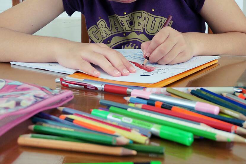 Een foto van een kleurboek en allerlei stiften, waarin een kind kleurt, we zien alleen diens armen en handen.