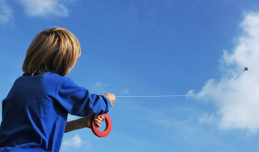 Een foto van een blauwe lucht, we zien de rug van een kind die vliegert