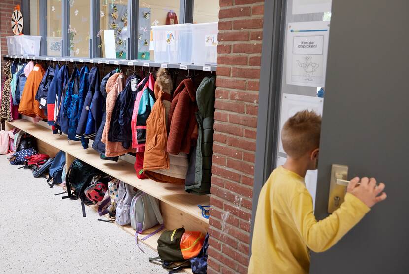Een kindje opent de deur van een klaslokaar. In de gang zie je jassen hangen en tassen staan op de grond