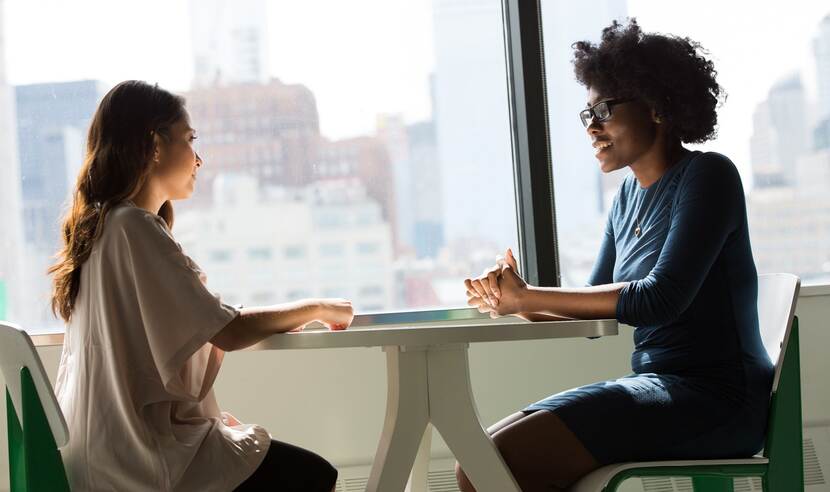 Een witte en een bruine vrouw zitten tegenover elkaar aan een ronde tafel. Ze zijn met elkaar in gesprek.