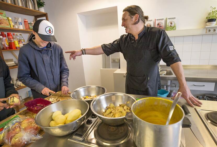 Een jongen snijdt lachend groente. Een man met een kookschort wijst lachend naar hem. Op het fornhuis staat een schaal aardappelen en een pan met eten