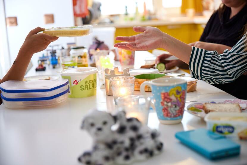 Een foto van een ontbijttafel, we zien dat een kinderhand een pakje kaas doorgeeft aan een volwassen hand